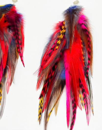  A pair of vibrant feather earrings displayed against a white background. The earrings are adorned with a variety of feathers in bold colors, including red, yellow, blue, and purple, with some natural brown feathers as well. Each earring has a silver fish hook style fastener with a small sunburst patterned charm attached to the top. The feathers are arranged in a cascading, slightly curved fashion, giving the earrings a dynamic and flowing appearance.