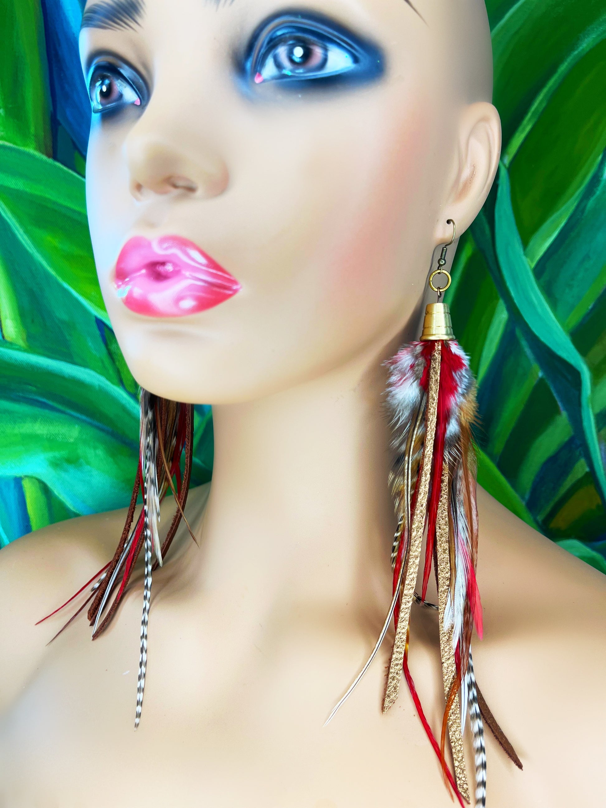 A mannequin wearing long feather earrings in brown and deep red, with metallic bronze leather strips, hanging from a gold colored cap pendant.