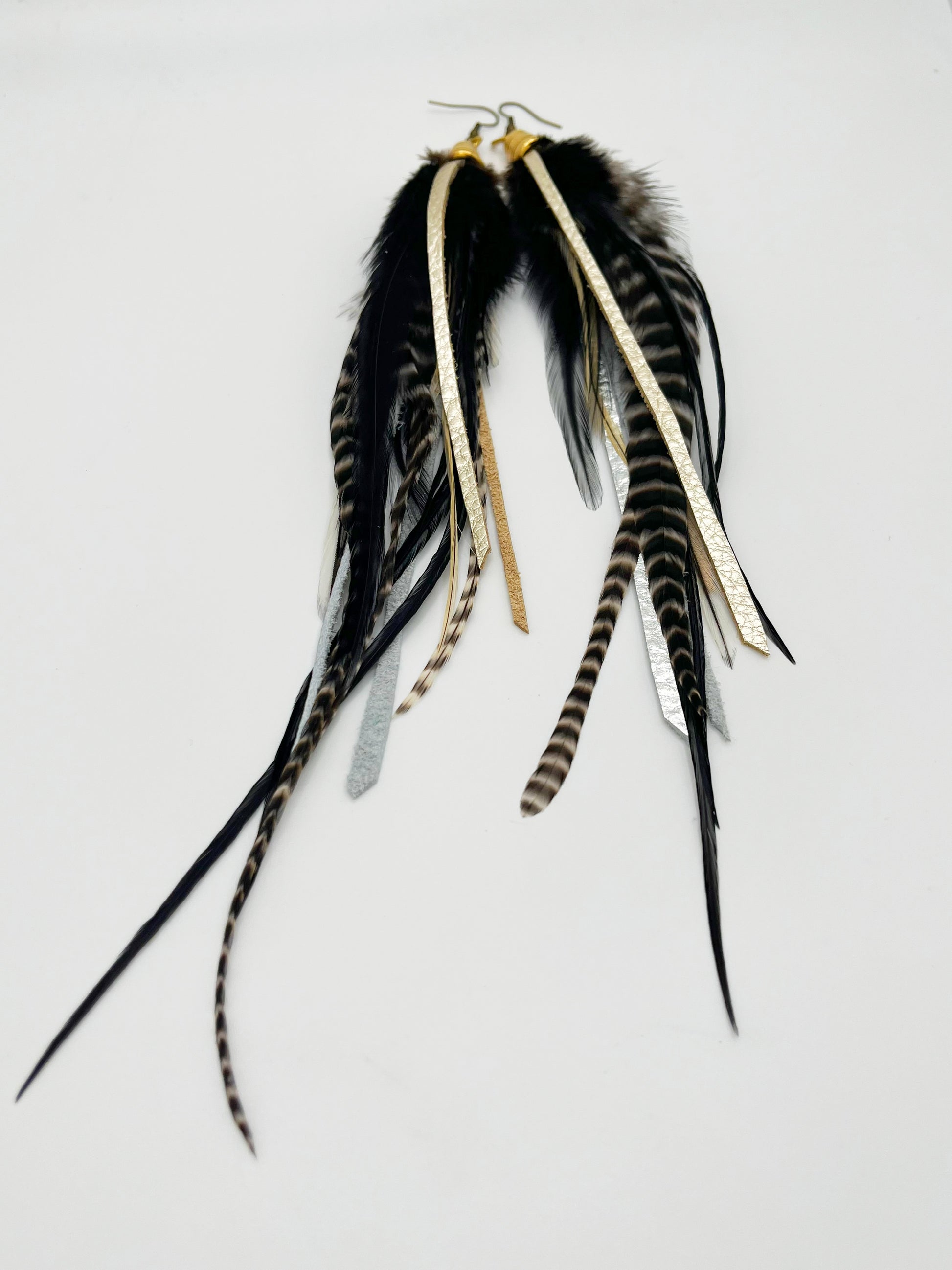 Long black and white feather earrings, with metallic silver and pewter colored leather strips, hanging from a gold colored cap pendant.