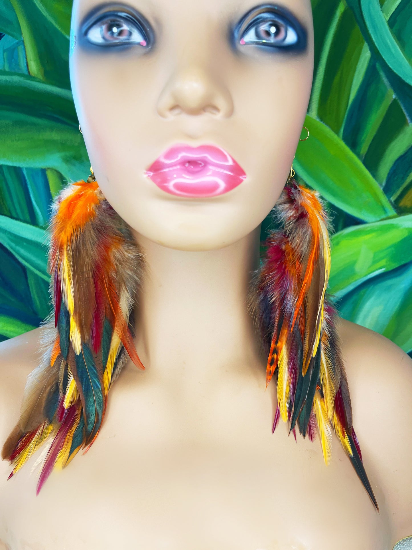 Mannequin showcasing 'Autumn's Gaze' Long Feather Earrings with a blend of orange, red, and yellow feathers and a gold link chain against a tropical leaf background.