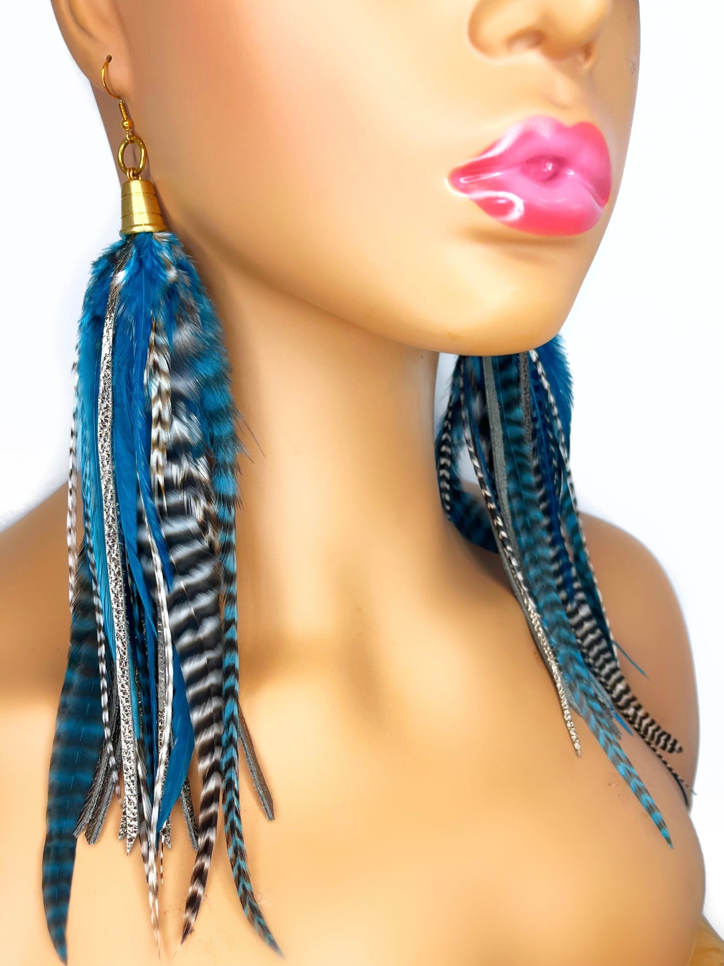 Mannequin wearing long teal rooster feather earrings with strips of metallic bronze leather. Feathers and leather hang from under a gold colored pendant.