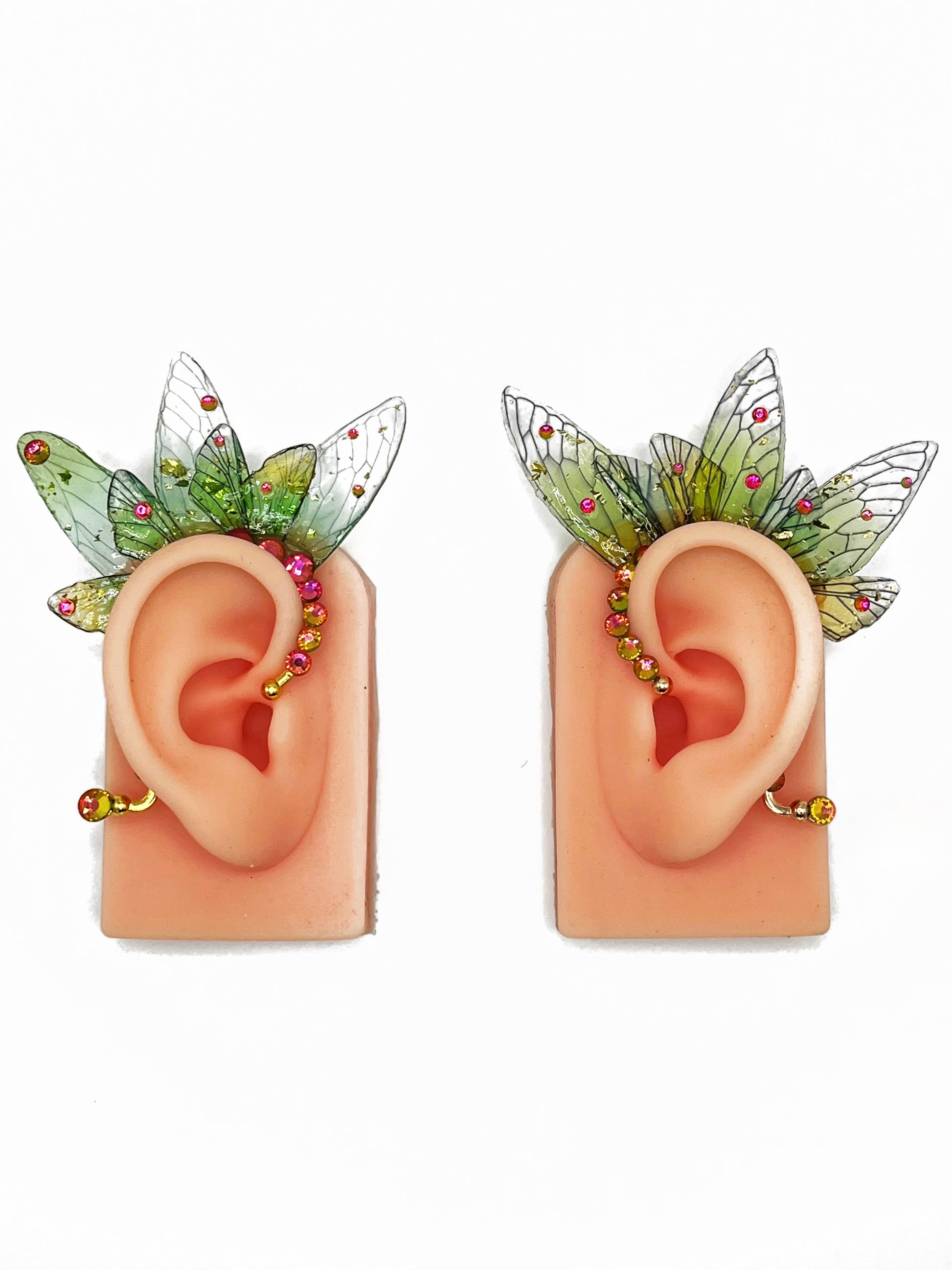 A pair of green fairy wing ear cuffs with pink rhinestones, modeled on a pair of ears. 