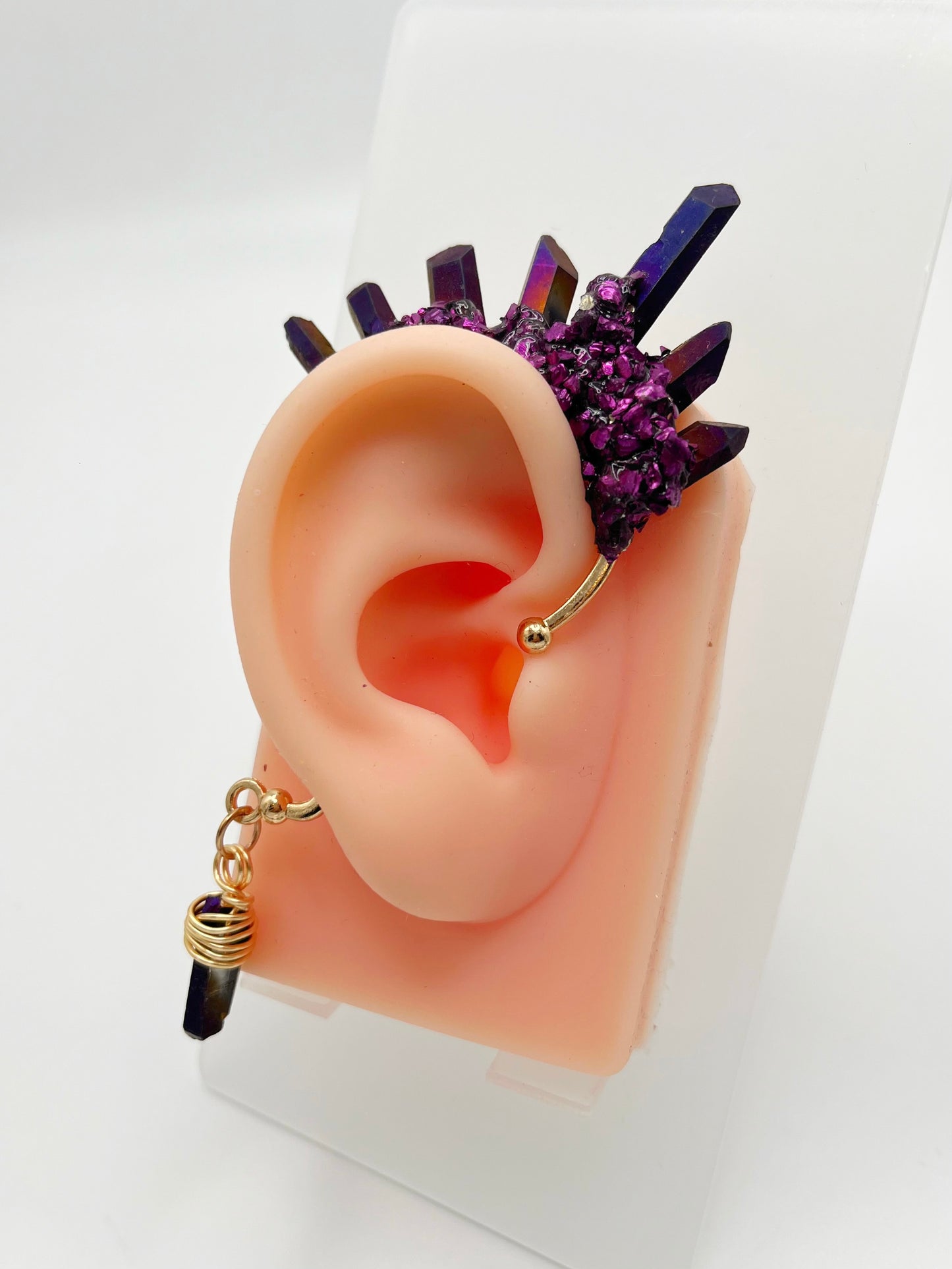 Purple titanium coated quartz ear cuff with metallic purple accents, modeled on an ear. A wire wrapped quartz stone is attached to the bottom of the cuff that hangs behind the ear.
