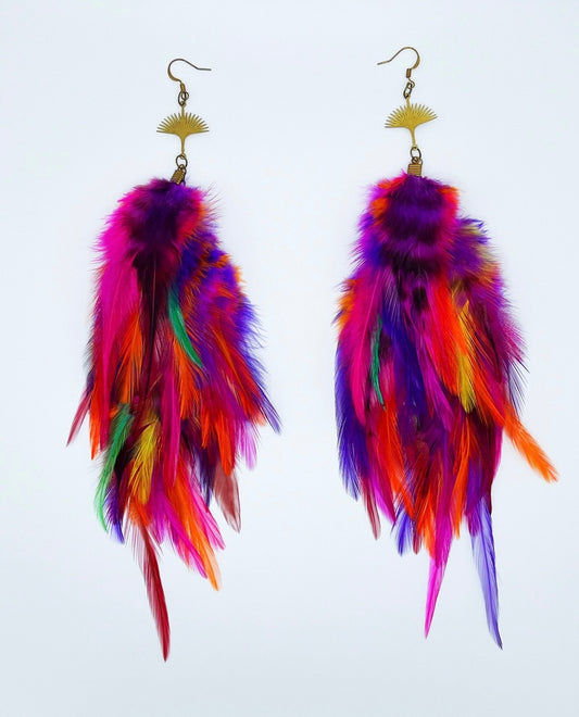 A pair of "Tropical Rave" Long Bohemian Feather Earrings displayed against a white background. These vibrant earrings feature a riot of colors with feathers in shades of purple, orange, pink, yellow, and green. They are connected to a gold fan-shaped pendant that attaches to a gold ear hook with a sunburst design. The feathers fall in a lush, layered pattern that creates a dynamic and full-bodied appearance, embodying the lively spirit of a tropical festival.