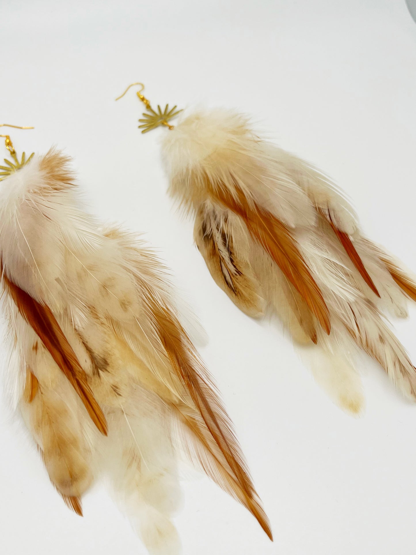 Long bohemian feather earrings in cream and beige tones, hanging from a gold sun pendant. 