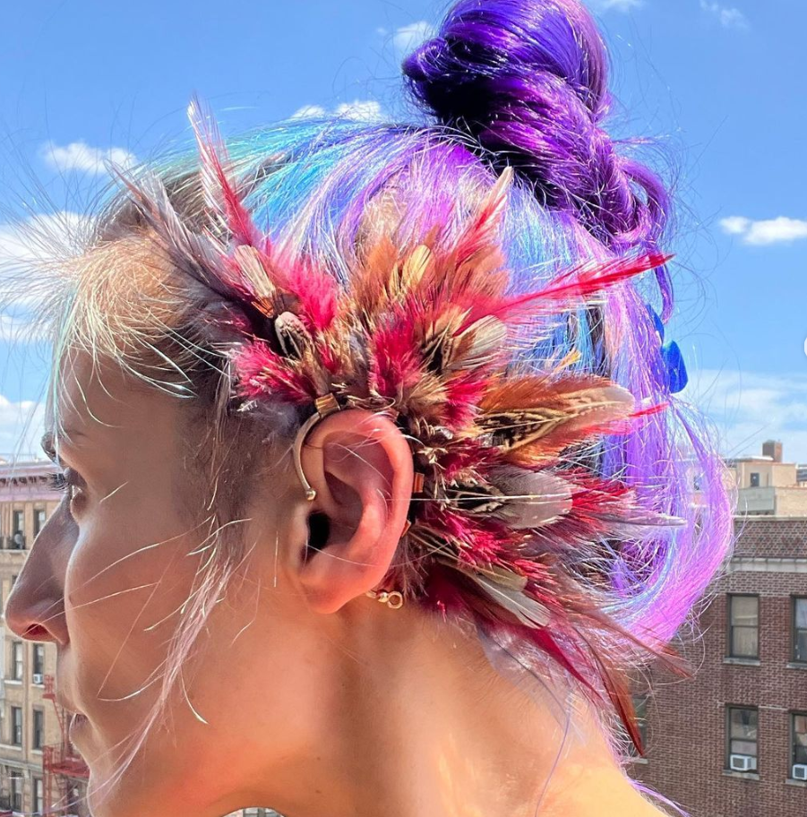 Side profile of a woman with vibrant purple hair and a colorful feather ear cuff, embracing a bold festival style.