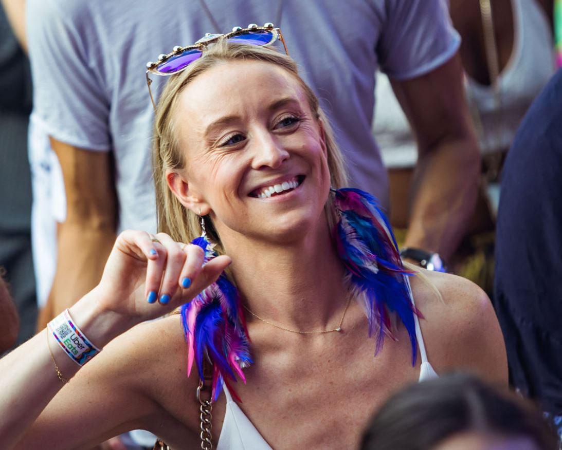 Woman smiling at a festival wearing vibrant blue and purple feather earrings and matching sunglasses on her head.